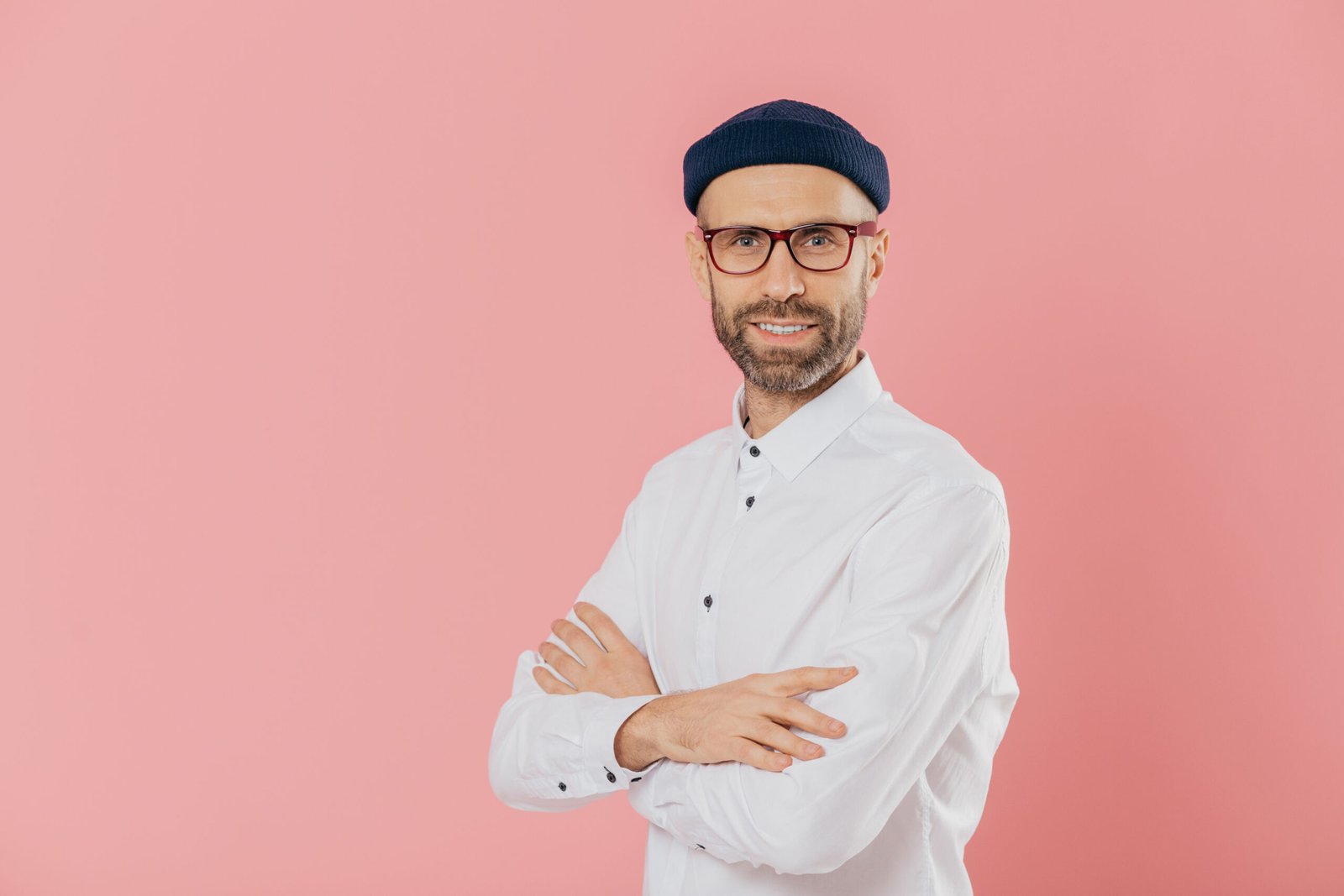 Positive male entrepreneur with self confident satisfied facial expression, keeps hands crossed, stands in profile, looks directly at camera, poses indoor against pink background with free space